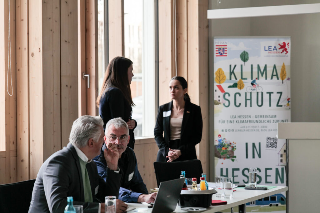 Eine Workshopszene wird gezeigt. Teilnehmende unterhalten sich in einem Veranstaltungsraum. Im Hintergrund ist ein Roll-Up mit dem Slogan zu sehen: "Klimaschutz in Hessen".