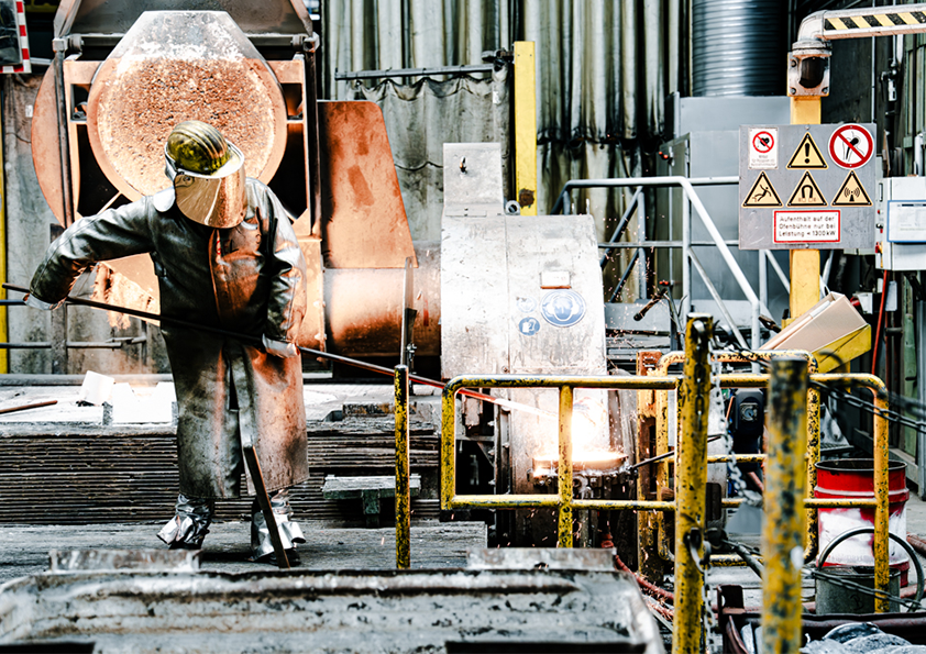 Das Bild zeigt die Produktionshalle der VAKUUMSCHMELZE. Ein Mitarbeiter ist in Hitzeschutzkleidung von Kopf bis Fuß gekleidet, während er mit heißem Metall hantiert.
