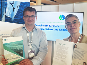 Caroline Werner und Rafael Noster stehen in Vertretung für die Geschäftsstelle am Informationsstand der Initiatve Energieeffizienz- und Klimaschutz-Netzwerke. Sie halten die Erfolgsgeschichten-Broschüre in ihren Händen.