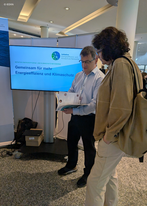 Rafael Noster steht am IEEKN-Informationsstand mit einer Interessentin und blättert die Erfolgsgeschichten-Broschüre durch.