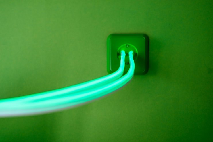 Eine grüne Steckdose auf grüner Wand, aus der grüner Strom in grellen Farben fliesst zur Versinnbidlichung von sauberer Energie.