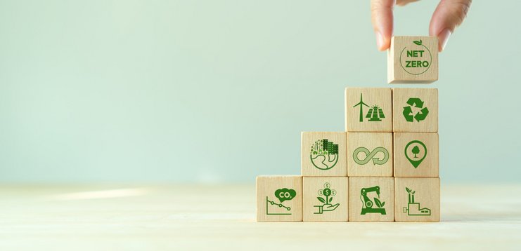 Eine Hand platziert aufeinander gestapelte Holzwürfel mit verschiedenen Icons zum Thema Nachhaltigkeit, Netto-Null und Klimaneutralität auf mintfarbenem Hintergrund.